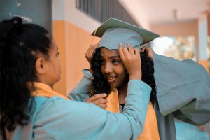girl in blue graduation gown with another girl helping her with mortarboard