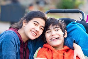 Big sister hugging disabled brother in wheelchair outdoors, smiling